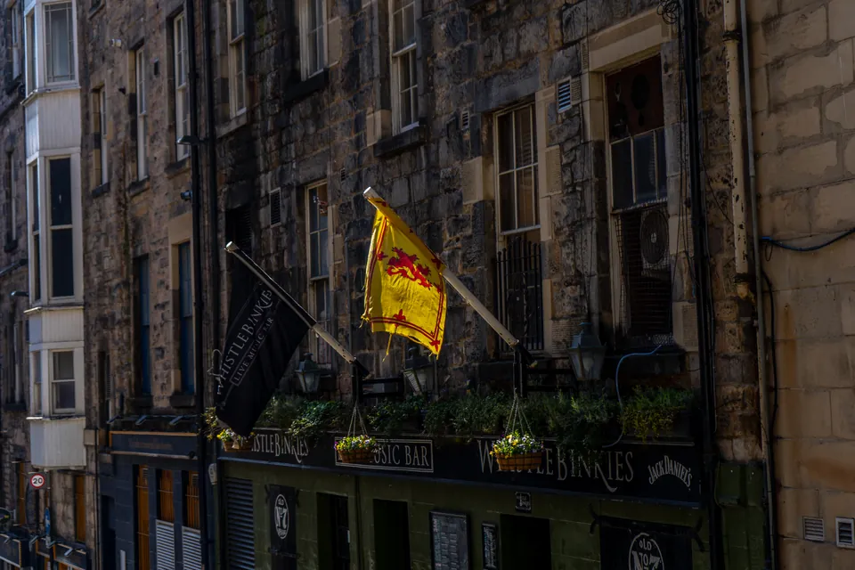 Sunlight catches a yellow Lion Rampant flag above an Edinburgh pub, against dark stone tenements