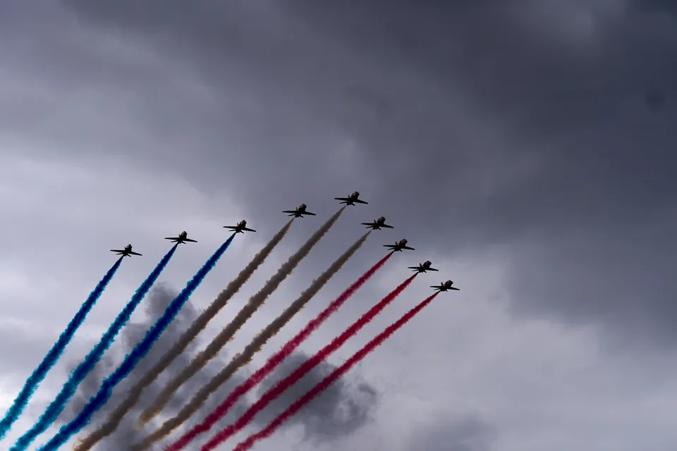 The Red Arrows climb in formation with parallel red, white, and blue smoke trails against a cloudy sky