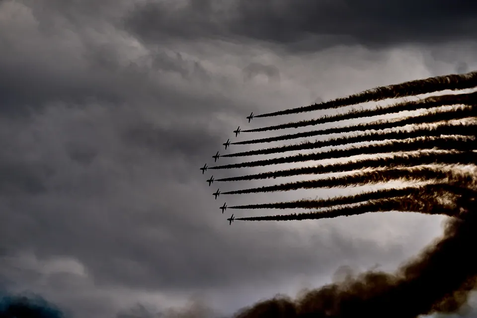 An aircraft formation curves through dark clouds above sweeping smoke trails