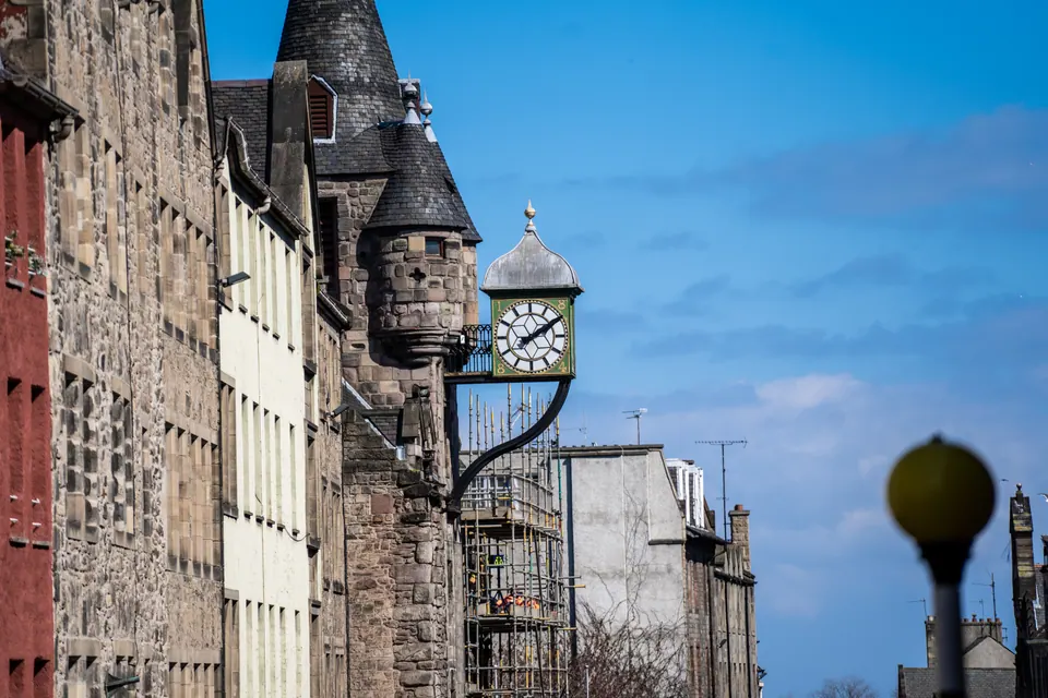 A projecting clock and turret rise above Edinburgh stone buildings against a blue sky.