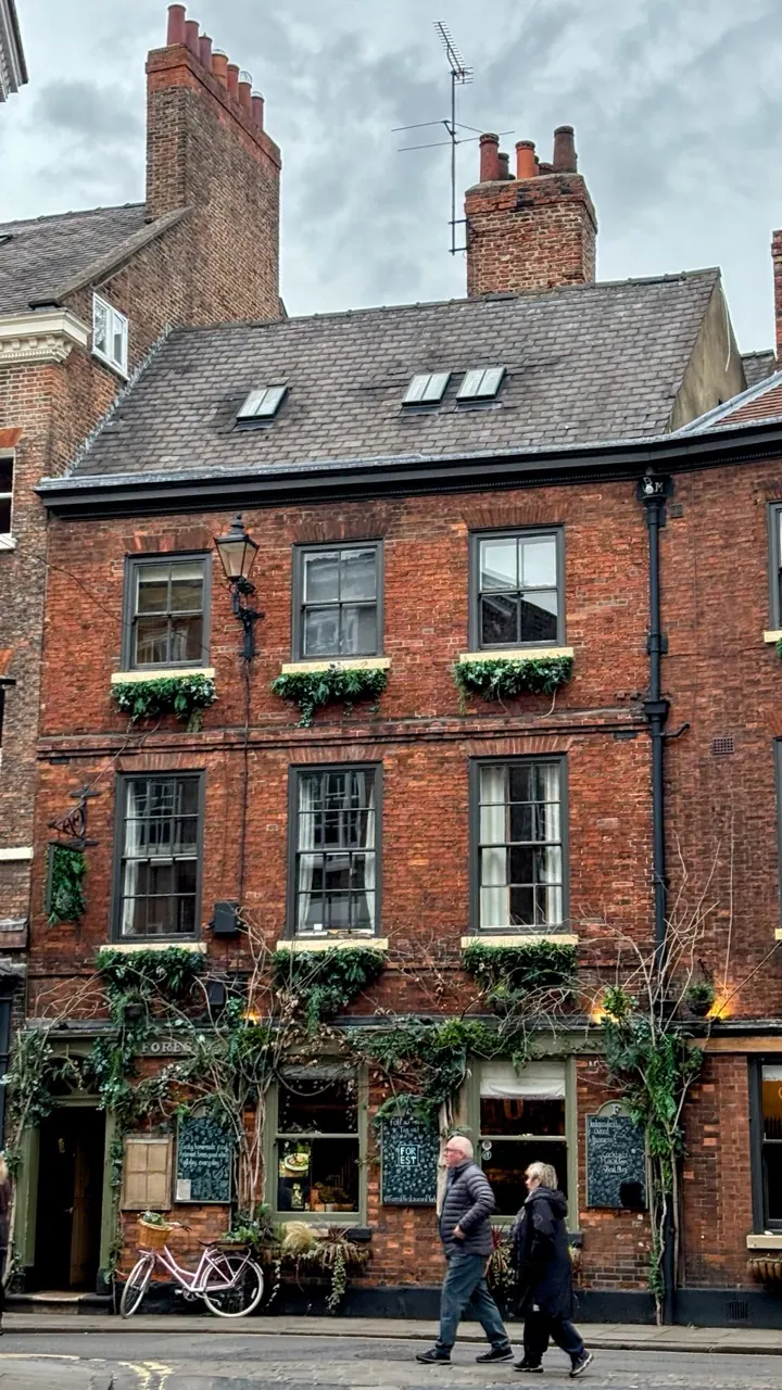 A red brick pub with hanging flowers and people passing on the pavement