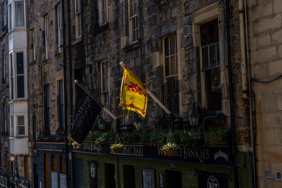 A yellow Lion Rampant flag above a dark green pub front, beneath Edinburgh stone tenements.