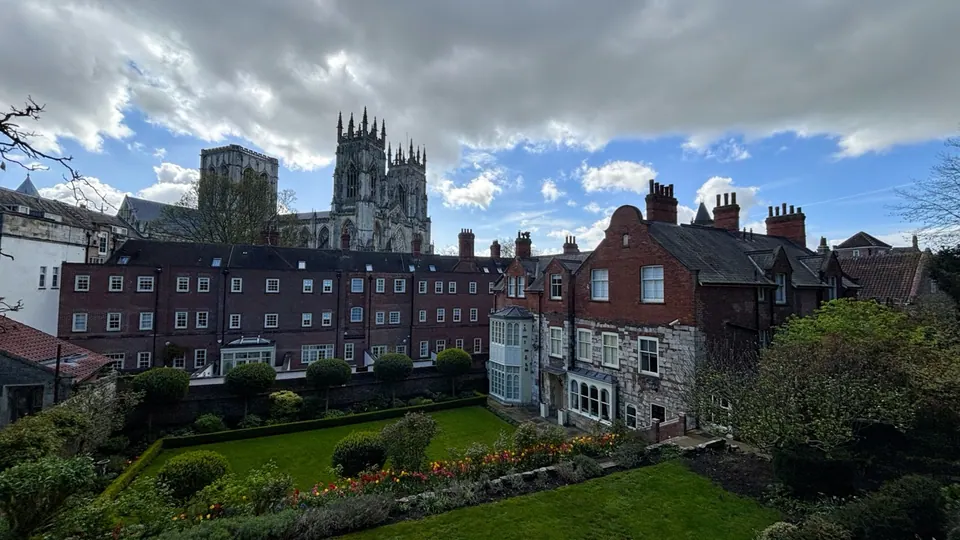 A cathedral rises above red-roofed buildings and a green garden beneath broken clouds