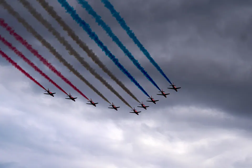 The Red Arrows descend across a cloudy sky with coloured smoke stretching towards the upper left
