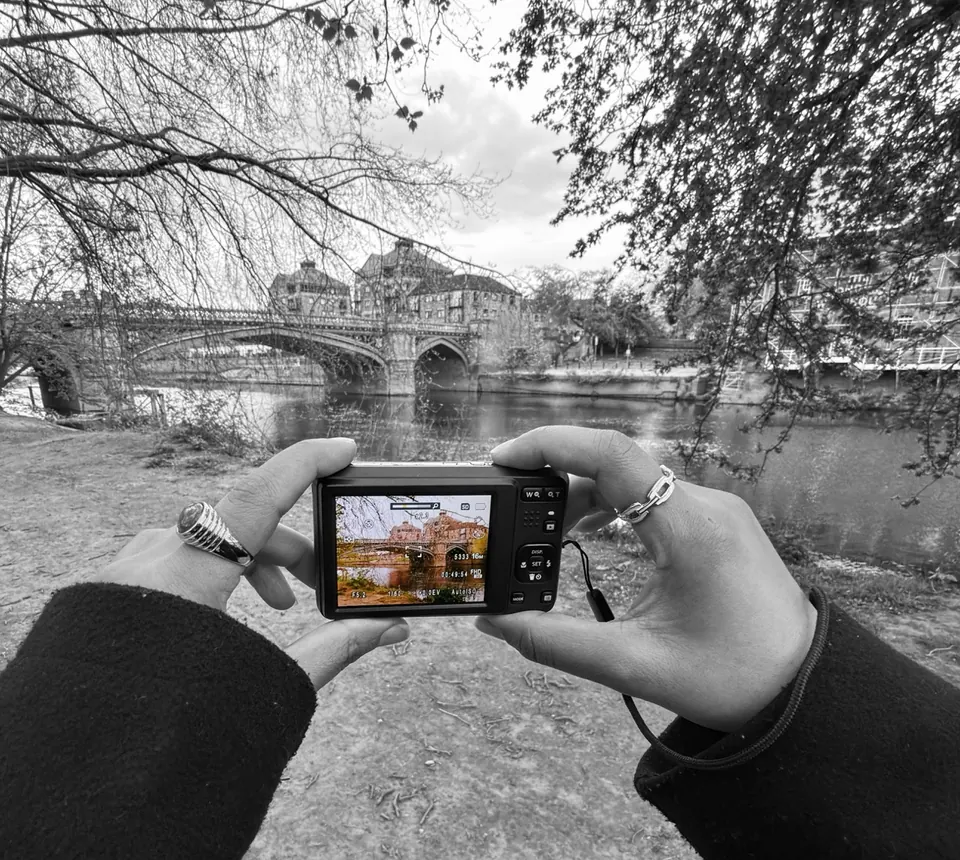 Hands frame a stone bridge on a compact camera, with the surrounding scene in black and white